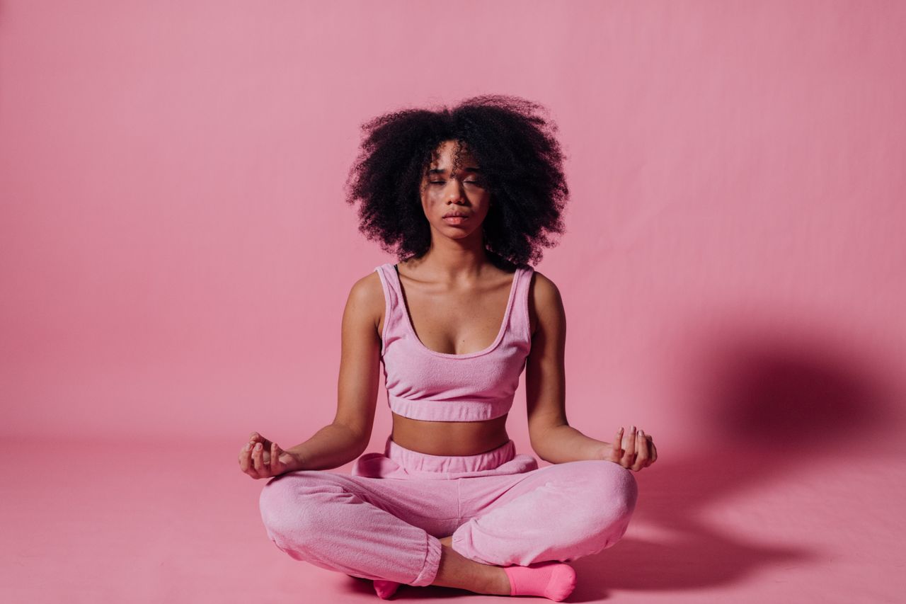 services-06 A woman with afro hair meditates in a peaceful studio with pink surroundings, embodying relaxation and zen.